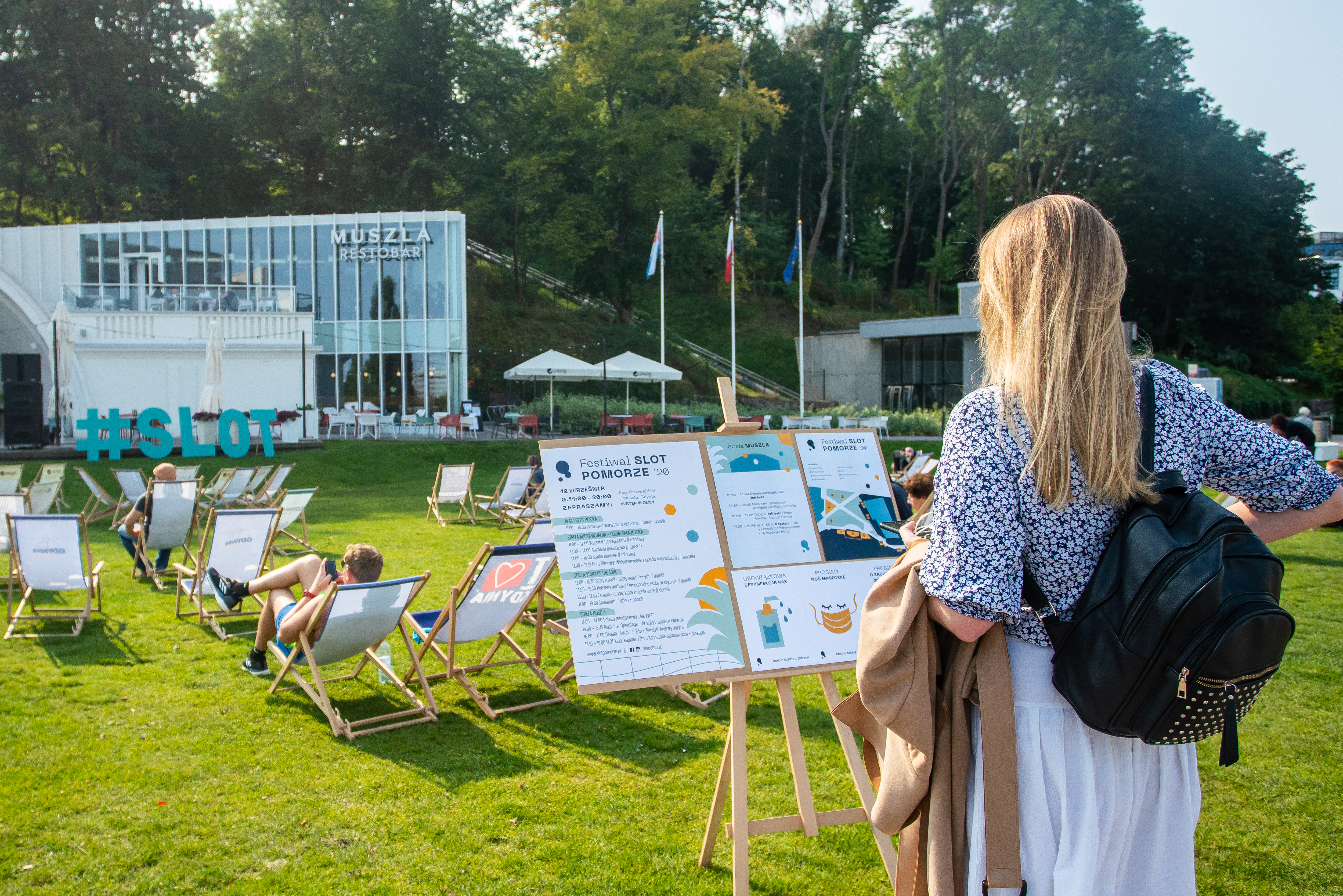 Jedna z uczestniczek przed tablicą z programem festiwalu SLOT Pomorze. W tle siedzący na leżakach ludzie // fot. Anna Rezulak