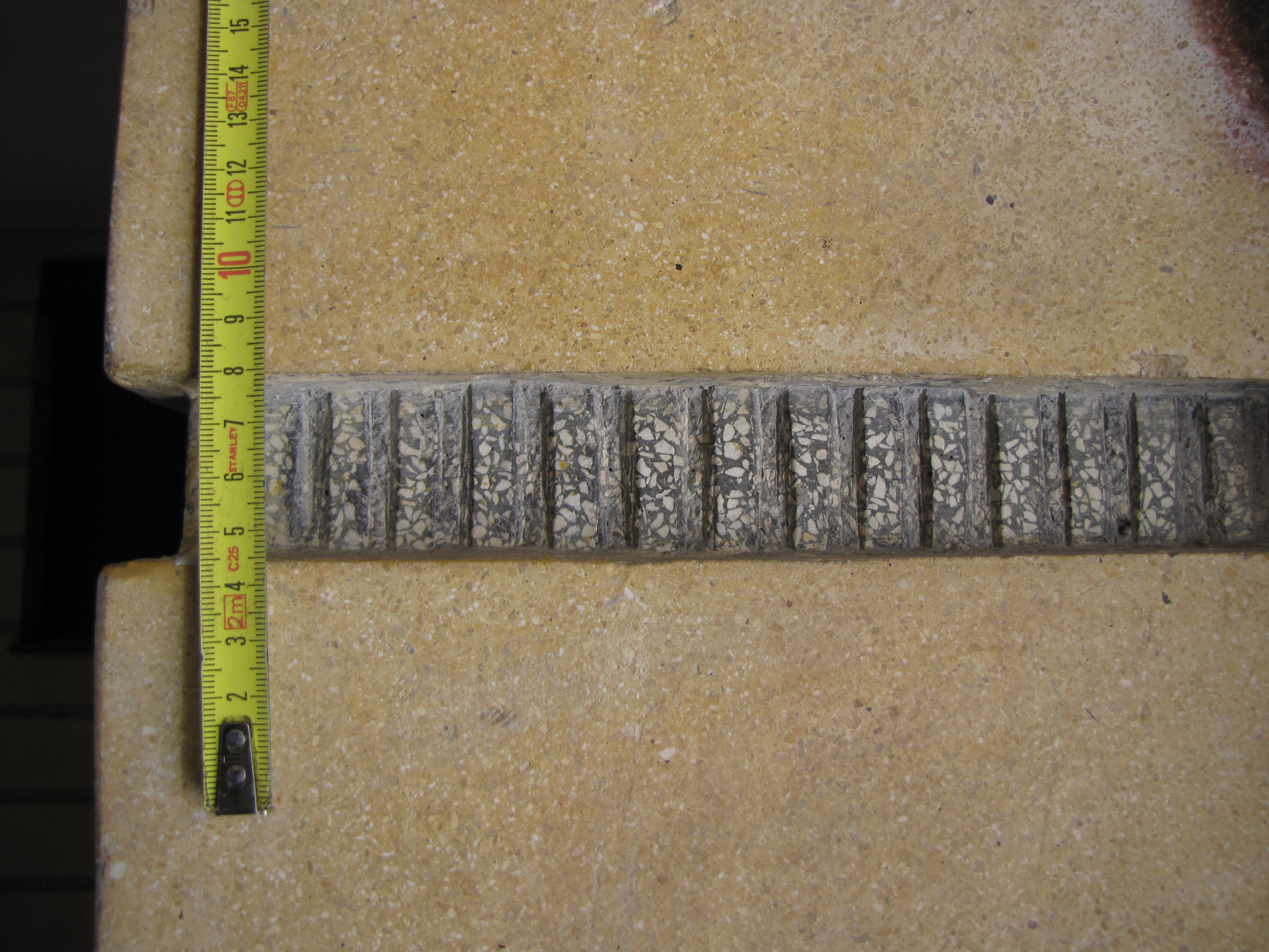 Fragment wykończenia tynkarskiego ściany podcienia budynku przy ul. 3 Maja 27-31 z pasami ż&oacute;łtego szlifowanego tynku rozdzielonego rowkami, w kt&oacute;rych wykonano tynk w kolorze grafitowym z białymi kamykami dodatkowo ponacinany pionowo