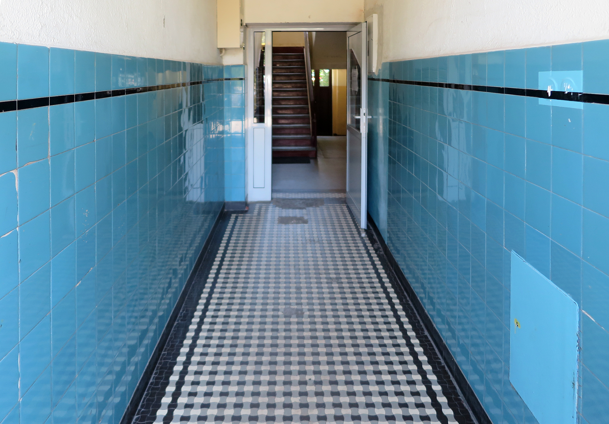 Strefa wejściowa w kamienicy przy ul. Portowej 3, dolne partie ścian wykończone ceramicznymi błękitnymi płytkami z wykończeniem z czarnym paskiem, posadzka zakomponowana z czarno-białych płytek tzw. gorsecik&oacute;w.