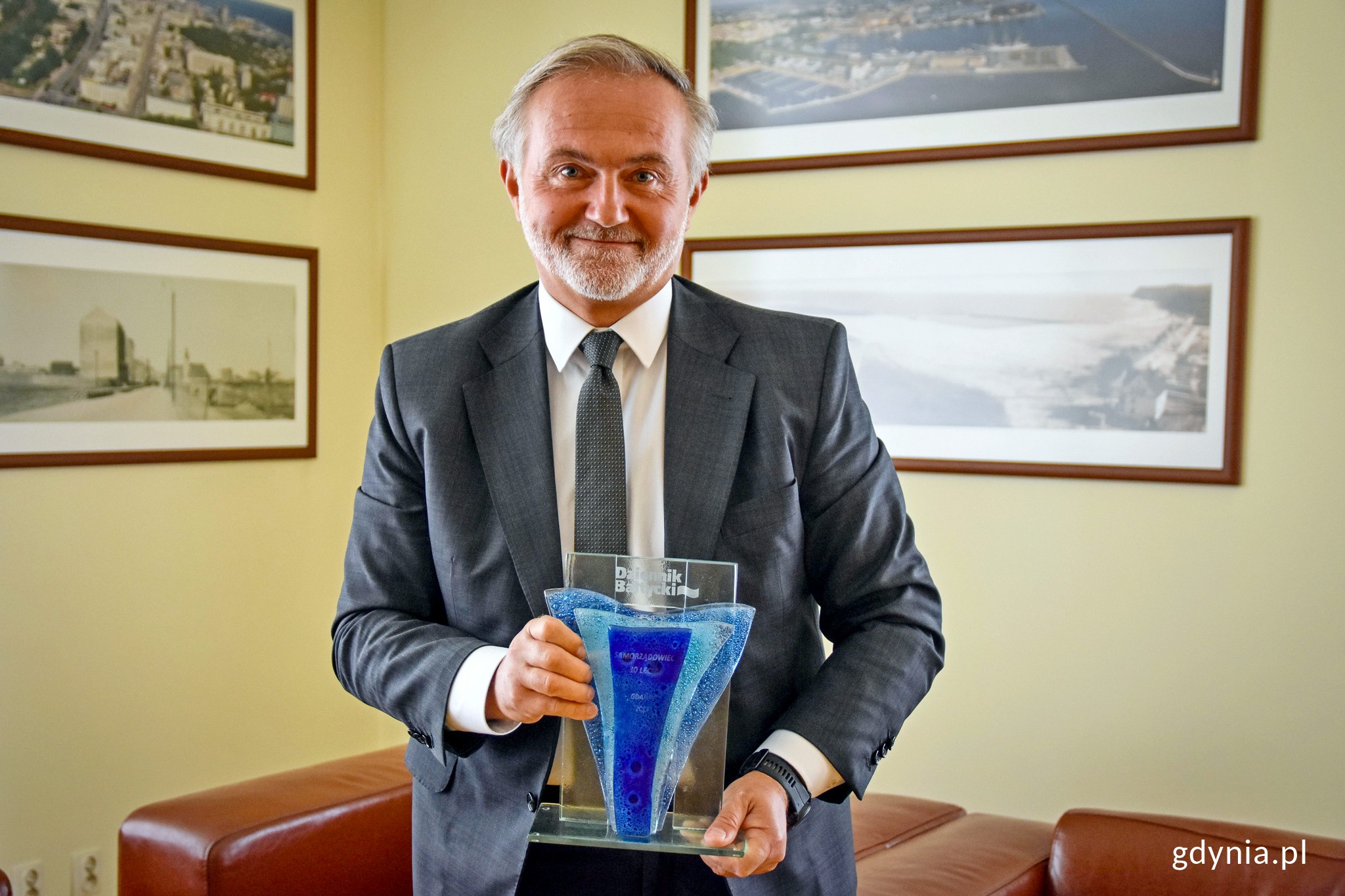 Prezydent Gdyni Wojciech Szczurek, ubrany w białą koszulę, grafitową marynarkę i krawat, trzyma w rękach nagrodę otrzymaną w konkursie &bdquo;Samorządowiec 30-lecia Dziennika Bałtyckiego&rdquo;. Nagroda jest wykonana z przezroczystych oraz niebieskich i błękitnych element&oacute;w. Widnieje na niej logo "Dziennik Bałtycki". W tle: biuro oraz fotografie przedstawiające dawną Gdynię.